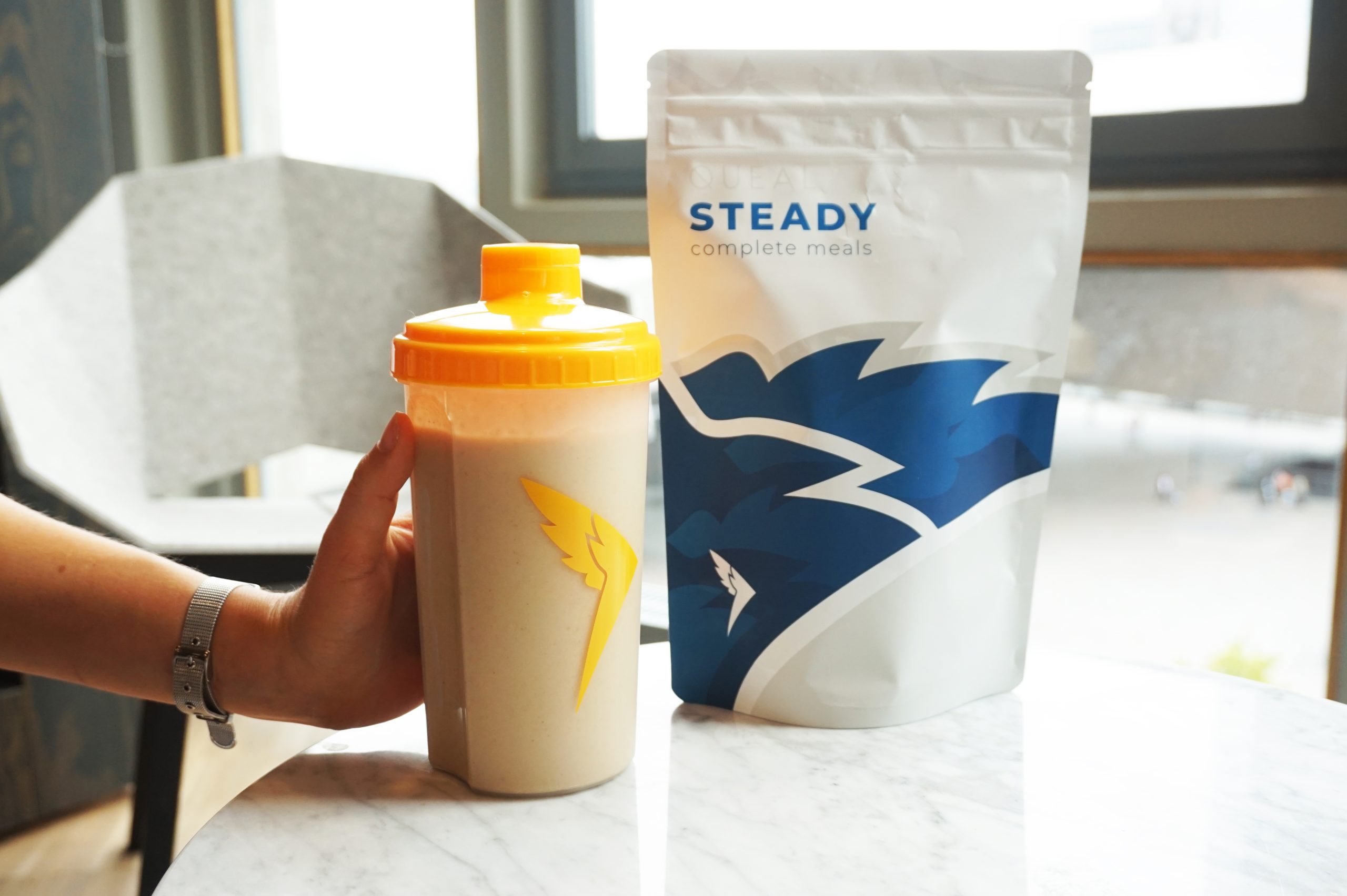 Classic Raspberry - Flow 2 A hand holding a Queal shaker with an orange lid, filled with a Queal shake, next to a pouch of Queal Steady complete meals, placed on a marble table with a window in the background.