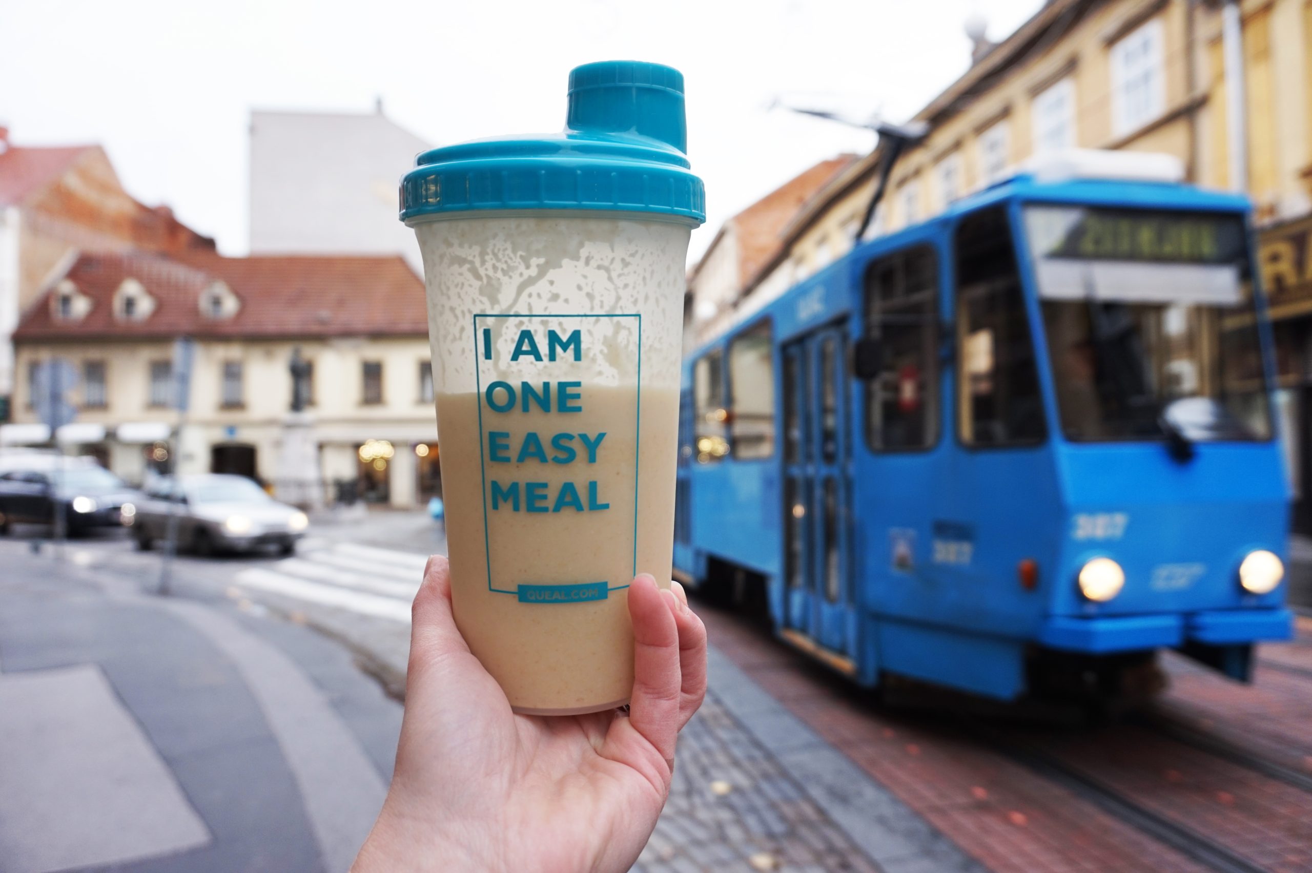 Classic Raspberry - Flow 3 A hand holding a Queal shaker with a blue lid, filled with a Queal shake, with the text "I AM ONE EASY MEAL" on the shaker, in front of a city street scene with a blue tram and buildings.
